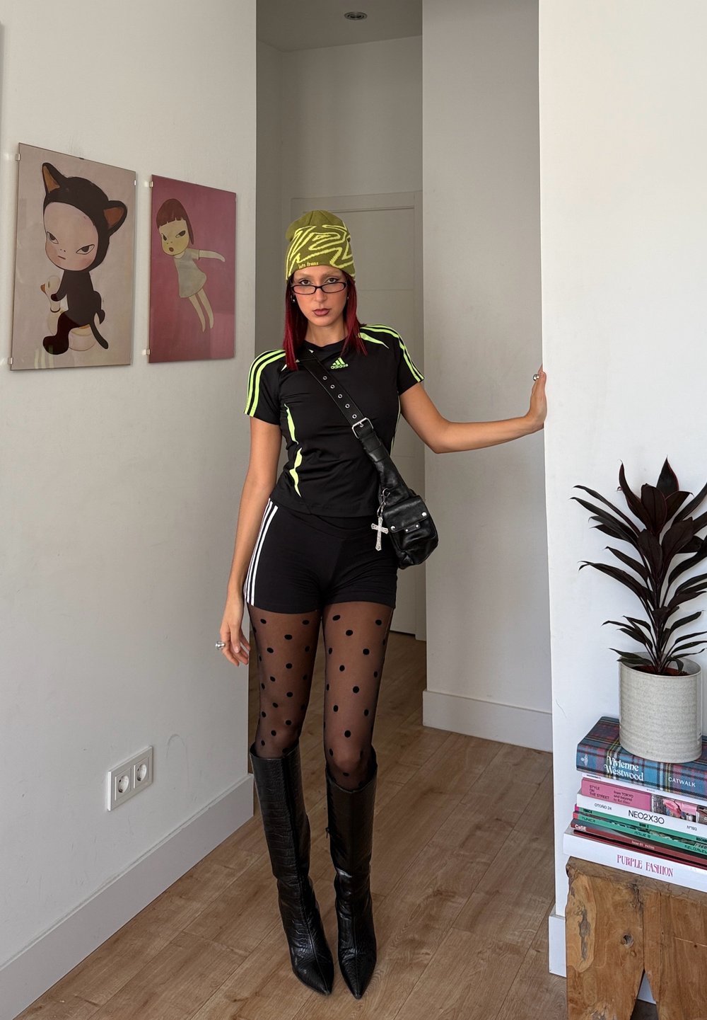 Joven mujer con atuendo atlético negro y mallas de lunares se encuentra en el interior junto a una pared blanca con arte moderno y una planta en maceta.