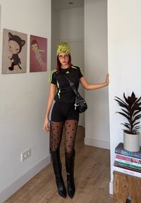 Joven mujer con atuendo atlético negro y mallas de lunares se encuentra en el interior junto a una pared blanca con arte moderno y una planta en maceta.