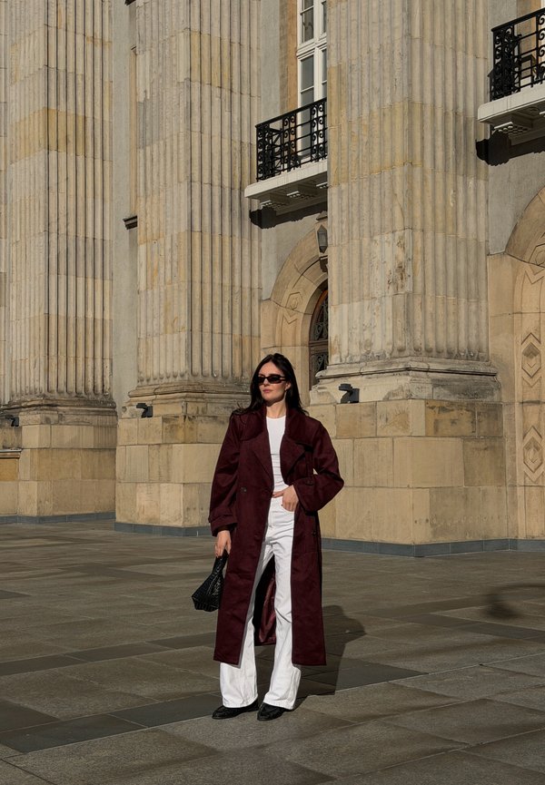 Mujer con un abrigo largo rojo oscuro, pantalones blancos y gafas de sol está junto a grandes columnas de piedra de un edificio histórico bajo la luz del sol.