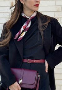 Femme portant un manteau noir et une robe avec une ceinture bordeaux, une écharpe fleurie, un sac à main bordeaux assorti, et du rouge à lèvres rouge, devant un mur en briques.