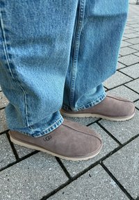Sapatos slip-on em camurça castanha com solas de borracha bege, apresentando detalhes costurados e um logótipo 'UGG' de lado, combinados com jeans azuis.