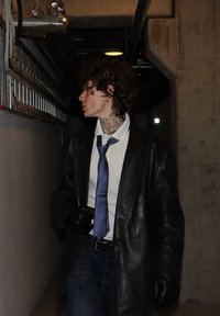 Czarny skórzany płaszcz na białej koszuli i niebieskim krawacie, ciemne jeansy oraz ręka w rękawiczce. Widoczne kręcone włosy i tatuaże na karku. Betonowe tło.