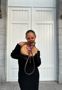 Woven rattan handtas met een roze accent en een bruine leren band, vastgehouden tegen een zwart geribd trui voor een witte paneeldeur.