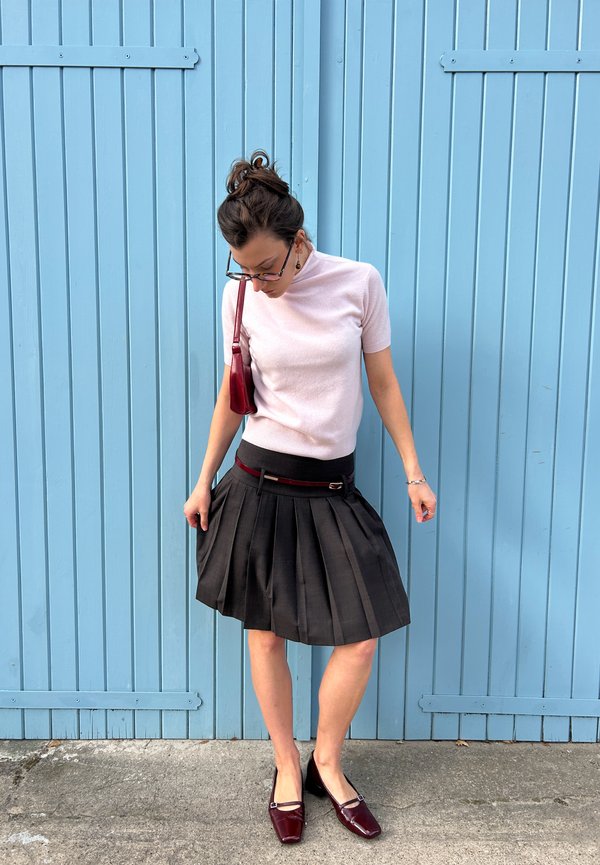 Top rosa a maniche corte abbinato a una gonna nera plissettata con una cintura rossa, completato da scarpe basse bordeaux e una piccola borsa rossa.
