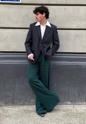 Blazer negro con rayas finas, camisa blanca de cuello y pantalones verdes de pierna ancha. Zapatos negros y auriculares inalámbricos blancos. Fondo urbano.