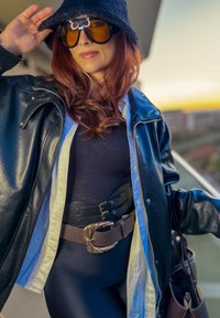 Chaqueta de cuero negra sobre una camiseta oscura, cinturón marrón con hebilla dorada en forma de media luna, gorro tipo pescador negro y gafas de sol grandes.