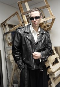 Veste de moto en cuir noir avec fermetures éclair et épaulettes, portée sur une chemise blanche, associée à une jupe noire. En arrière-plan, des caisses en bois.