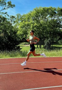 Bėgikas, apsirengęs smėlio spalvos sportiniu marškinėliu ir juodomis šortais, sėkmingai bėga raudonu takeliu. Jam ant kojų baltos sportinės bateliai ir akyse saulės akiniai, o fone matyti žali medžiai.