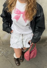 T-shirt bianco con grafica a fiocco rosa, giacca di pelle nera, gonna bianca con cerniere, stivali marroni fino al ginocchio e borsa rosa.