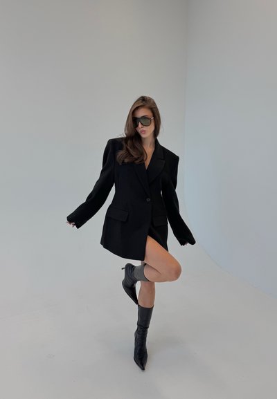 Blazer negro de gran tamaño con hombros estructurados y dos bolsillos frontales, llevado sobre piernas desnudas y combinado con botas altas hasta la rodilla con punta afilada.