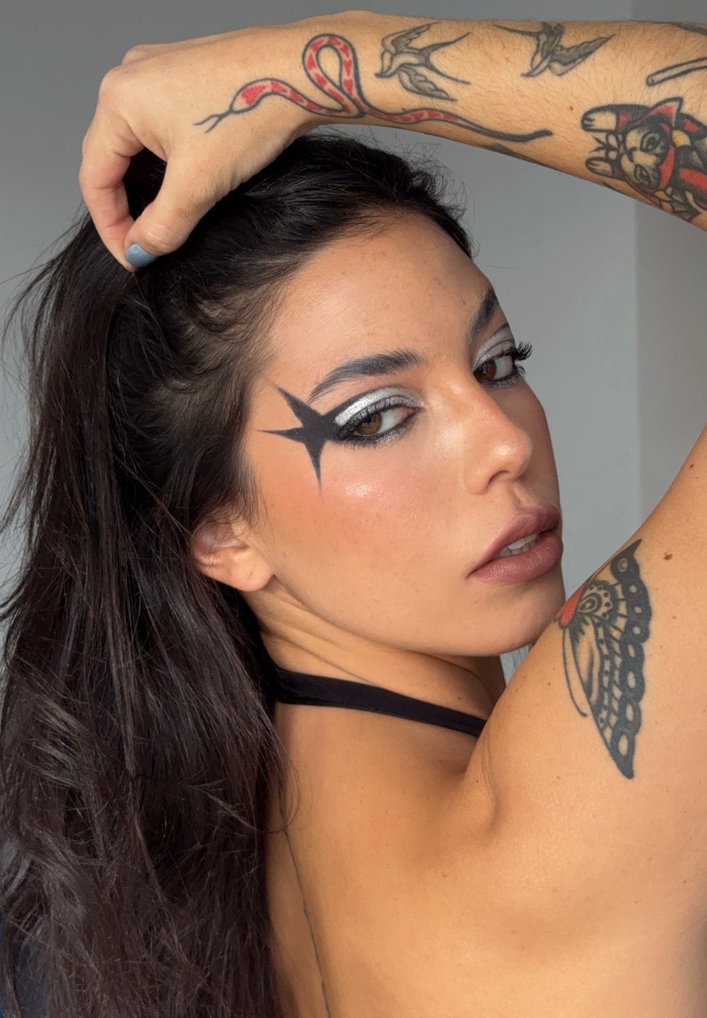 Svart stjärnsmink på vänstra sidan av ansiktet ovanför silver eyeliner, tillsammans med mörkt hår och arm tatueringar med detaljerade mönster.