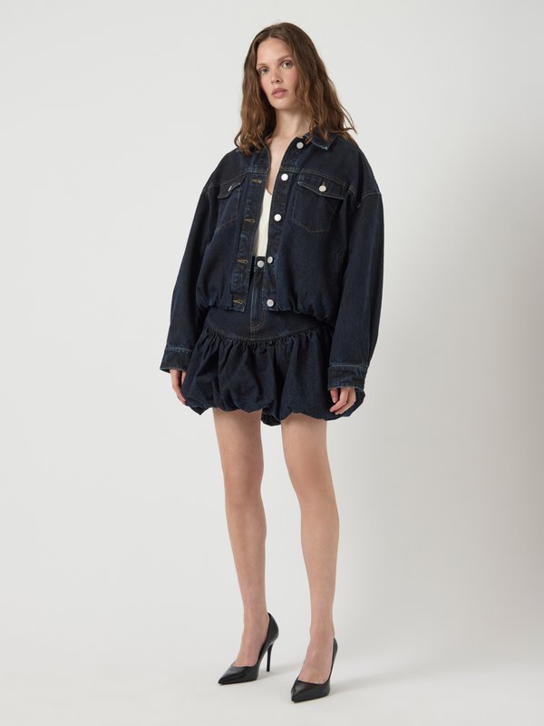 Τζάκετ από τζιν με oversized εφαρμογή, κλείσιμο με κουμπιά και δύο μπροστινές τσέπες, συνδυασμένο με φούστα από τζιν με φουσκωτό σχέδιο. Μαύρες ψηλές γόβες ολοκληρώνουν το σύνολο.