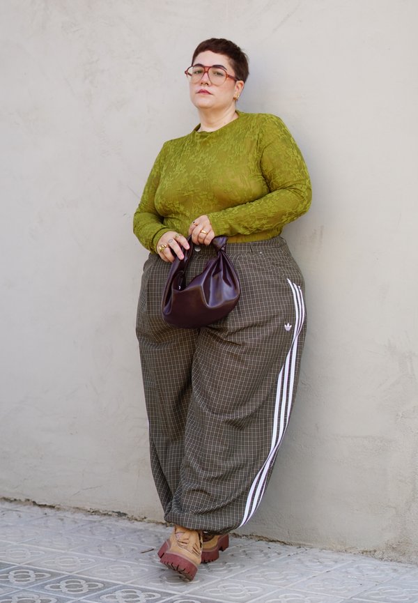 Persona con capelli corti che indossa un top verde strutturato, pantaloni a quadri con strisce bianche, stivali beige e tiene una piccola borsa scura, appoggiata a un muro.