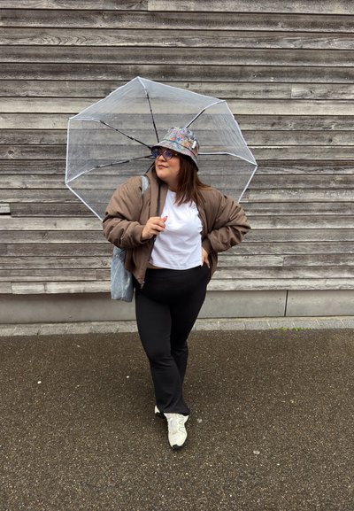 Paraguas transparente sobre una persona con una chaqueta bomber marrón, camiseta blanca, mallas negras y un sombrero de cubo con estampado, de pie sobre un pavimento mojado.
