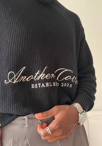 Persoon draagt een zwarte gebreide trui met de tekst "Another Cotton Established 2023", is een grijze broek aan het vastmaken, met een zilveren horloge en ring zichtbaar.