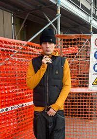 Melna polsterēta veste pār spilgti oranžu garroku kreklu, melna cepure; stāv pret oranžu sietveida barjeru. Redzamas tumšas džinsa bikses.