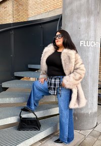 Personne portant des lunettes de soleil, un manteau en fourrure, un haut noir, un jean large et une ceinture-sac à carreaux, debout sur des escaliers en métal avec un sac à main noir sous le pied.