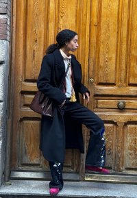 Zwarte jas, geborduurde zwarte broek met kleurrijk bloemenpatroon, witte shirt, en roze schoenen. Bruine tas en houten deur op de achtergrond.