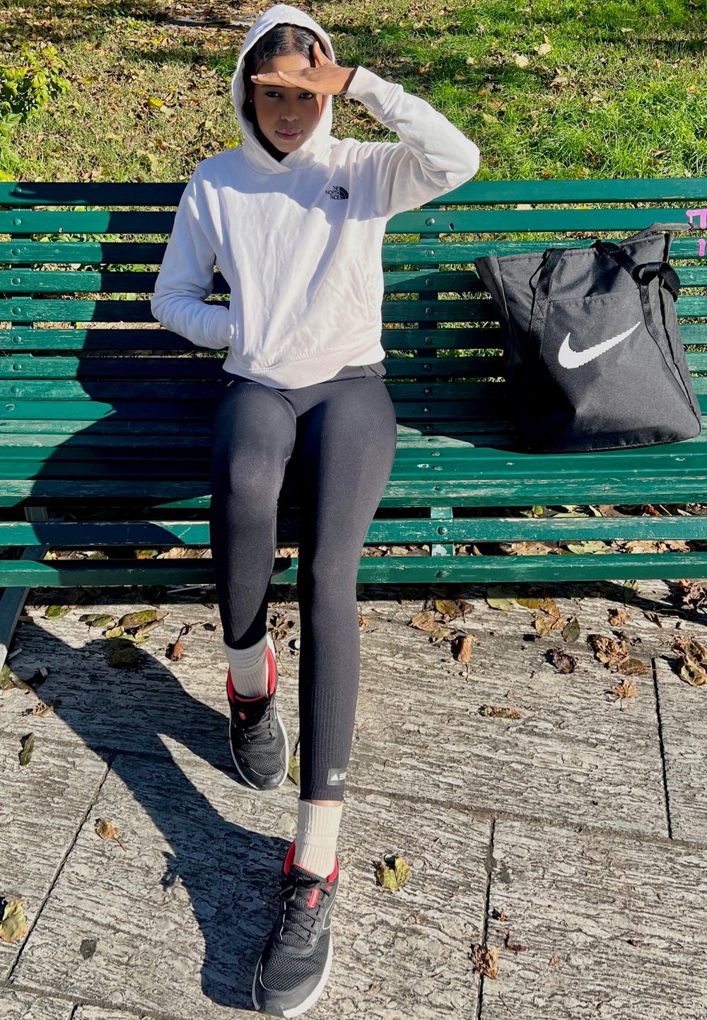 Hanorac alb cu glugă, colanți negri și adidași negri cu accente roșii. O geantă tote Nike neagră stă alături pe o bancă verde.