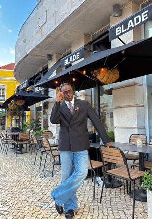 Hombre con blazer negro, corbata estampada y gafas, apoyado en una mesa de cafetería al aire libre bajo un toldo negro con letreros que dicen "BLADE".