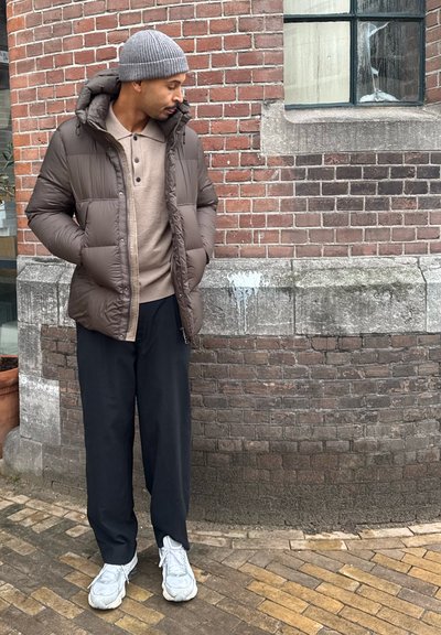 Chaqueta acolchada marrón con aislamiento y capucha, llevada sobre una camisa beige abotonada. Pantalones negros de pierna ancha y zapatillas deportivas gris claro completan el conjunto.