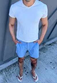 Camisa branca de algodão com decote em V, calções azul claro com bolsos laterais e chinelos brancos. O ambiente é uma superfície de betão ao ar livre.