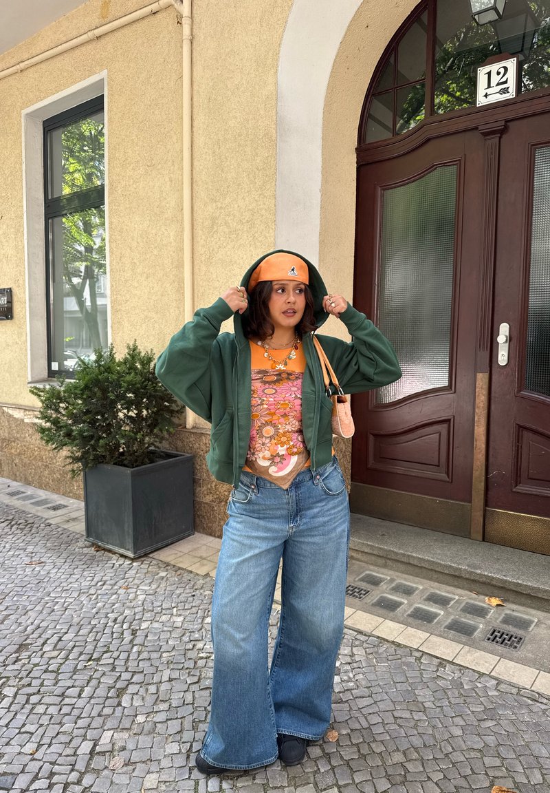 Felpa verde oversize, t-shirt grafica arancione, jeans a gamba larga blu e un berretto arancione abbinato. Gli accessori includono una piccola borsa a tracolla e una collana.