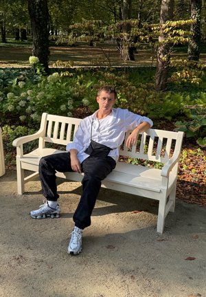 Jongeman met een bril, witte shirt, zwarte broek en sneakers, zittend op een wit bankje in een zonnig park met groene bladeren.