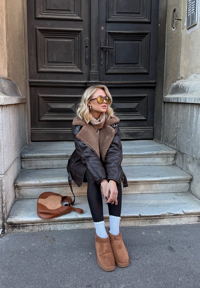 Casaco castanho forrado com shearling e gola bege, leggings pretas e botas de cano curto em camurça bege. Acessórios incluem óculos de sol amarelos e uma mala castanha.