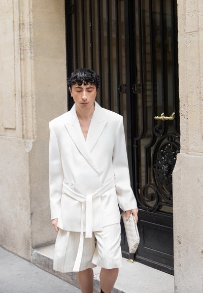 Persona con cabello corto y rizado lleva una americana blanca con cinturón y pantalones cortos plisados a juego, sosteniendo un bolso de mano tejido, de pie junto a una puerta de hierro forjado negra.