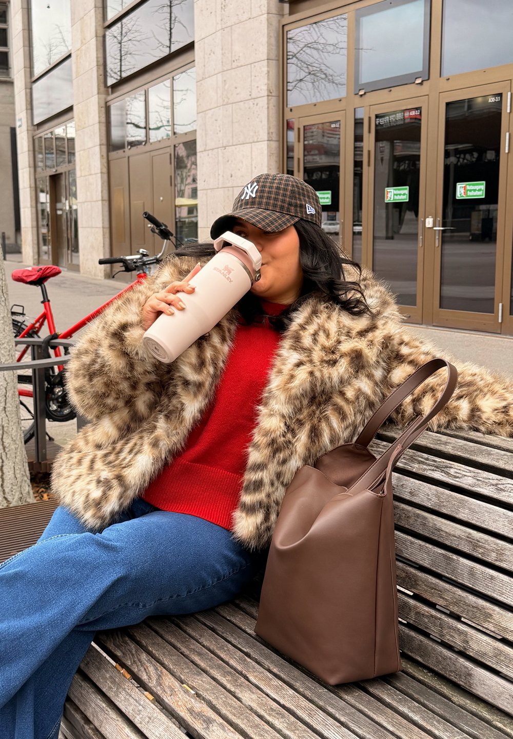 Manteau en fausse fourrure marron, pull en maille rouge, jeans évasés en denim bleu. Tient une bouteille d'eau isolante rose. Sac fourre-tout marron sur le banc.