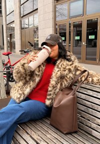 Manteau en fausse fourrure marron, pull en maille rouge, jeans évasés en denim bleu. Tient une bouteille d'eau isolante rose. Sac fourre-tout marron sur le banc.