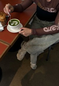 Ένα πράσινο ρόφημα με τέχνη latte σε ένα λευκό φλιτζάνι σε ένα πιάτο, δίπλα σε ένα τραγανό αρτοσκευάσμα, πάνω σε ένα κόκκινο τραπέζι. Το πρόσωπο φοράει ένα καφέ πουκάμισο με σχέδια και γκρι παντελόνι.