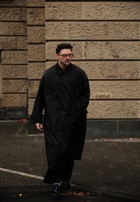 Must pikk mantel on lihtsa disainiga, nööpide kinnitusega, suure kraega ja lõdva istuvusega. Isik kannab musti kingi ja seisab tänaval.