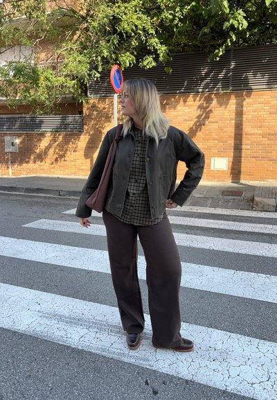 Chaqueta verde oscura, camisa de cuadros, pantalones de pierna ancha marrones, sosteniendo una bolsa marrón. En un paso de peatones con pavimento de color claro y un muro de ladrillos detrás.
