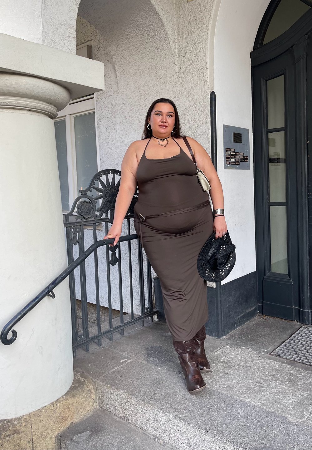 Καφέ, εφαρμοστό φόρεμα με λαιμόκοψη halter και απαλή υφή, σε συνδυασμό με καφέ, γόνατους, χαλαρούς μπότες. Τα αξεσουάρ περιλαμβάνουν μια μικρή τσάντα και ένα μαύρο καπέλο.