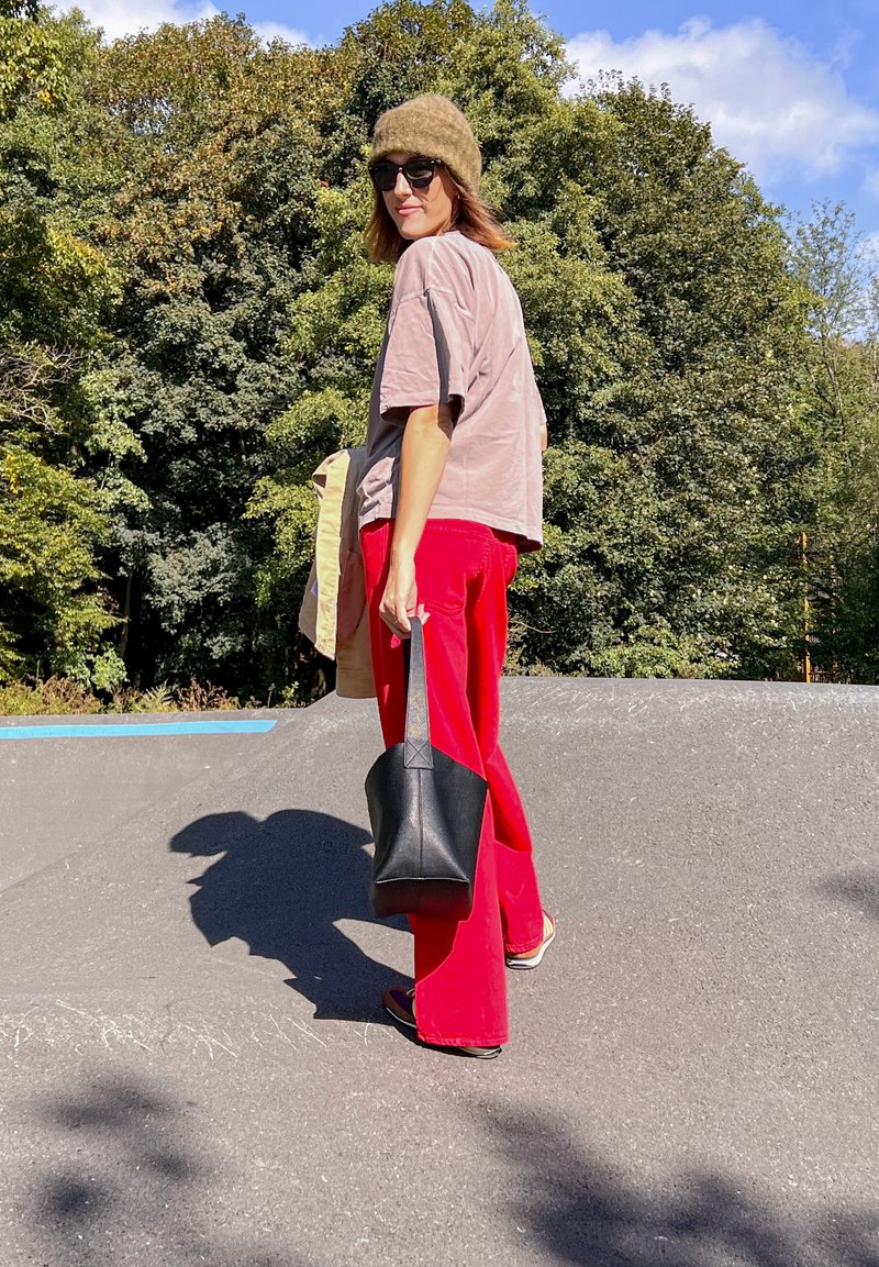Μοντέλο με ανοιχτό καφέ oversized πουκάμισο και φωτεινές κόκκινες παντελόνες, κρατώντας μια μαύρη τσάντα, με καφέ πλεκτό σκουφί και γυαλιά ηλίου.