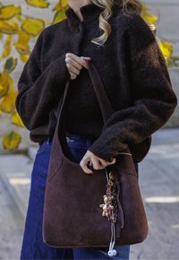 Pruun sametist õlakott, millel on sile tekstuur, kaardus kuju ja dekoratiivne ripats. Sobitatud tumeda kootud kampsuni ja siniste teksadega.