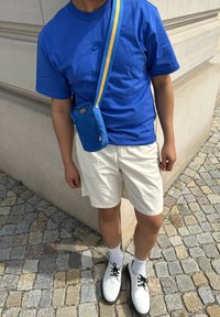 Maglietta Nike blu, pantaloni corti bianchi, scarpe bianche con lacci, borsa blu con tracolla gialla; in piedi su una superficie di ciottoli.