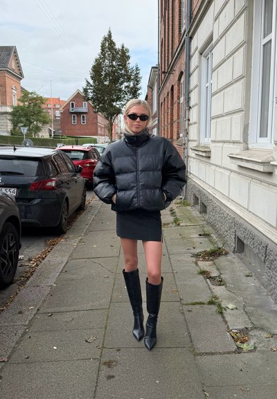 Chaqueta de plumas negra, ajustada, con un cuello alto. Llevada sobre una minifalda negra y botas negras hasta la rodilla. Corte entallado, textura suave.