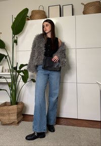 Femme debout à l'intérieur portant une veste grise en fausse fourrure, un haut noir sans manches, un jean à jambes larges, des chaussures noires et tenant un sac noir.