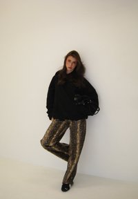 Sweat-shirt noir surdimensionné, pantalon brillant à motif serpent doré, et chaussures noires. Sac à main simple tenu contre la hanche, fond clair.