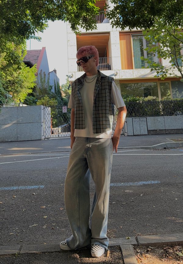 Personne aux cheveux roses et lunettes de soleil portant un gilet à carreaux, une chemise blanche, un jean large et des chaussures à motifs à carreaux, debout dans une rue devant des bâtiments.
