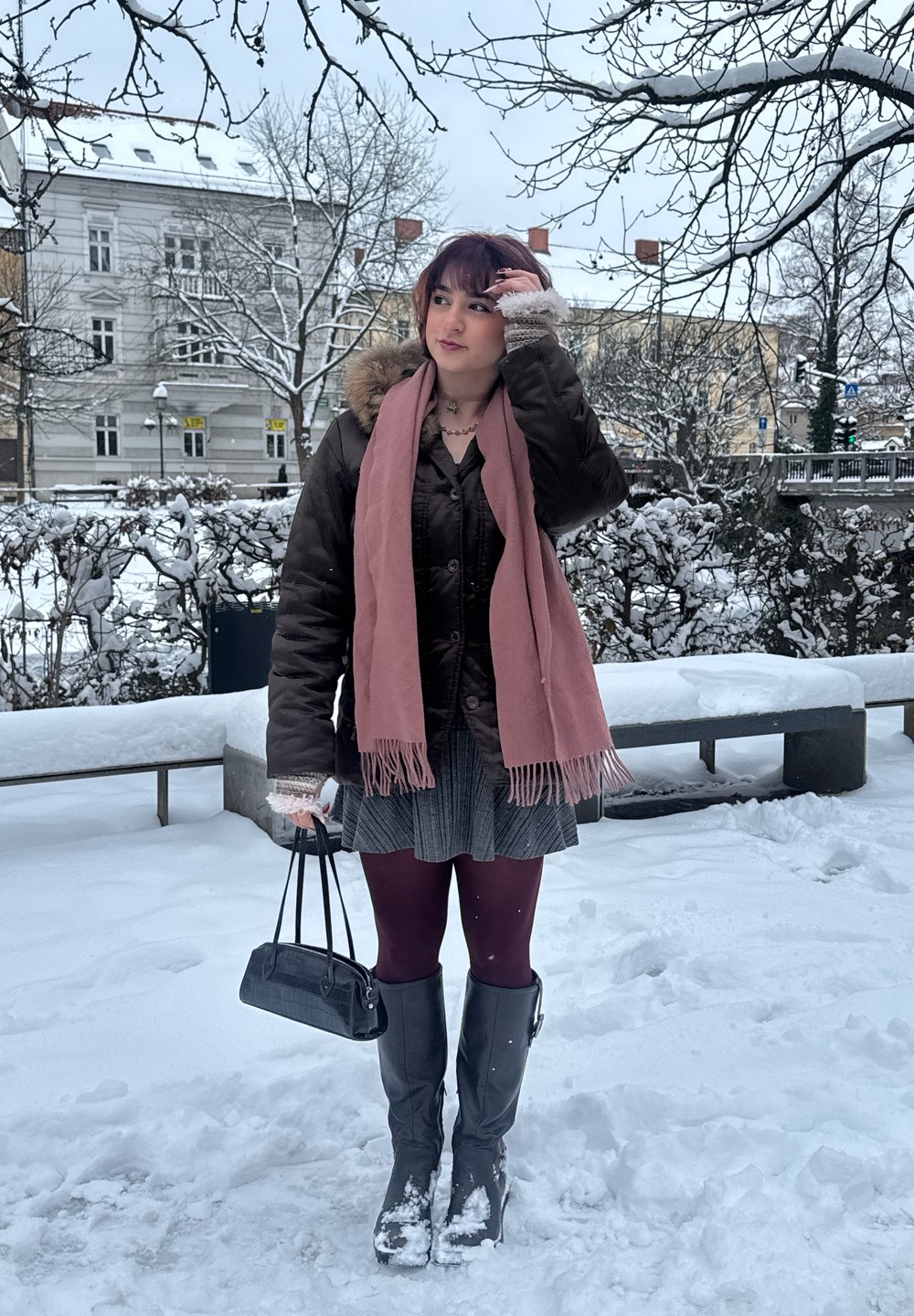 Doudoune marron avec capuche bordée de fourrure, écharpe rose, sac à main noir, jupe grise, collants bordeaux et hautes bottes noires dans un décor enneigé.