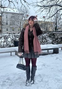 Doudoune marron avec capuche bordée de fourrure, écharpe rose, sac à main noir, jupe grise, collants bordeaux et hautes bottes noires dans un décor enneigé.