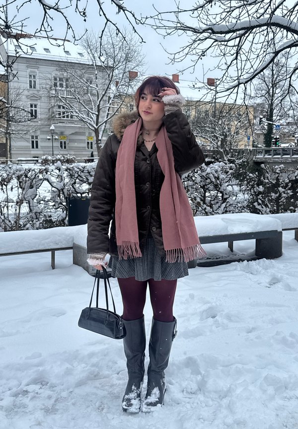 Rjava puhovka s kapuco obloženo z umetnim krznom, roza šal, črna torbica, siva krilo, bordo nogavice in visoki črni škornji v zasneženem okolju.