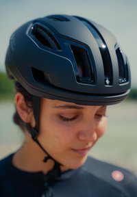 Capacete de ciclismo preto com aberturas de ventilação, acabamento mate e correias ajustáveis, usado por uma pessoa com uma camisola escura.