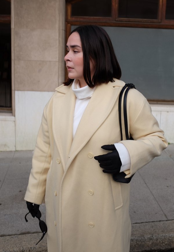 Donna con cappotto beige e guanti neri che tiene occhiali da sole e una borsa nera, guarda a sinistra all'aperto vicino a un edificio.