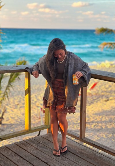 Mujer con un chal gris y una falda marrón sujetando una lata, de pie en una plataforma de madera con vistas a una playa de arena y un océano azul.