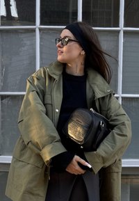 Olivgrüne übergroße Jacke, schwarze strukturierte Tasche mit goldenen Akzenten, schwarzer Pullover und graue Hose. Haare nach hinten gebunden, klare Brille getragen.